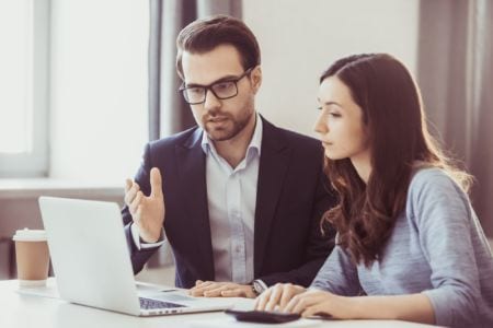 A man wearing glasses talking to a woman and looking at a laptop