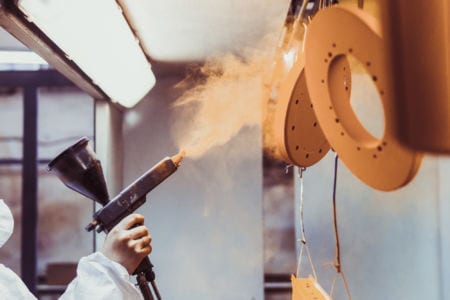 A worker in a protective suit spraying powder paint from a gun on a metal product